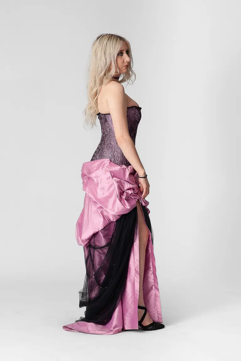 A woman stands in profile, wearing an elegant dress that features a purple bodice and a flowing, pink skirt with a black underlayer. The dress has a dramatic design with ruching, and she has long, wavy blonde hair. The background is neutral and plain.