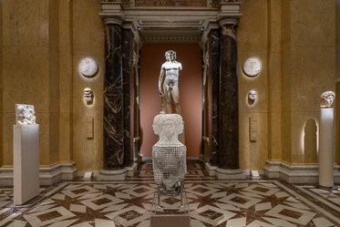Eine Museumsansicht mit einer weißen Statue des David in der Mitte. Um ihn herum sind verschiedene Skulpturen und Reliefs an den Wänden und auf Podesten ausgestellt. Der Boden ist mit einem auffälligen Mosaikmuster gestaltet. Die Wände sind aus warmem, goldenem Marmor.
