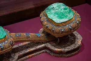 Eine kunstvoll gestaltete, goldene Feder mit einem großen, grünen Jade-Stein und bunten, eingelegten Edelsteinen. Sie liegt auf einem filigranen, reich verzierten Untergrund, der die exquisite Handwerkskunst und das kulturelle Erbe widerspiegelt.