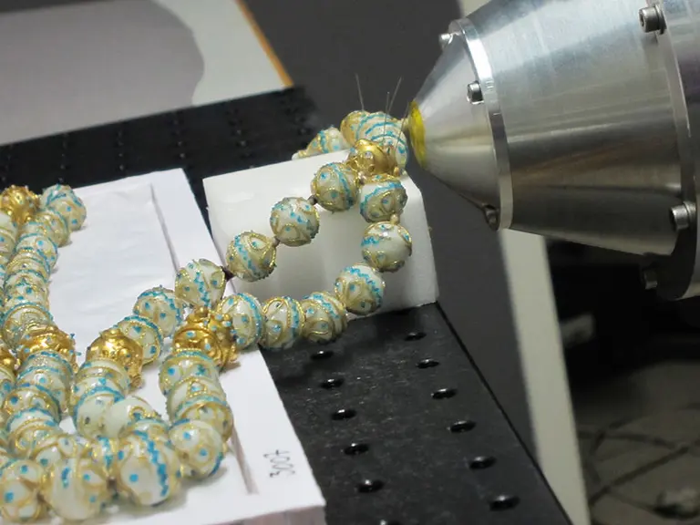 Ein Schmuckstück aus goldenen und bunten Perlen wird von einer Maschine bearbeitet. Die Perlen sind filigran gestaltet und liegen auf einem weißen Schaumstoffuntergrund. Die Maschine hat eine metallische Düse, die auf den Schmuck zielt.