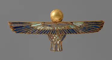 Ein goldenes Amulett in Form eines heraldischen Vogels mit weit ausgebreiteten Flügeln. Der Vogel hat einen runden, goldenen Kopf und ist mit blauen und grünen Emailleverzierungen geschmückt. Der Hintergrund ist grau.
