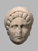 Eine antike Steinskulptur eines Frauenkopfes mit detaillierter Frisur und gleichmäßigen Gesichtszügen. Die Augen sind offen, und der Ausdruck wirkt neutral. Die Oberfläche ist leicht abgenutzt, was auf ihr Alter hinweist.