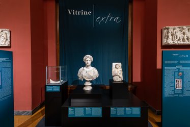 Eine Vitrine in einem Museum zeigt Skulpturen und Informationen. In der Mitte steht eine Büste, flankiert von zwei weiteren Objekten. Im Hintergrund hängt ein dunkelblaues Tuch mit der Aufschrift "Vitrine extra". An der Vitrine sind informative Plakate angebracht.