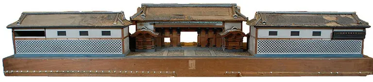 Dieses Bild zeigt ein detailliertes Modell eines traditionellen japanischen Gebäudes mit zwei seitlichen Strukturteilen und einem zentralen Tor. Die Wände sind mit einem charakteristischen Muster verziert, und das Dach ist leicht geneigt. Das Modell vermittelt einen Eindruck japanischer Architektur.