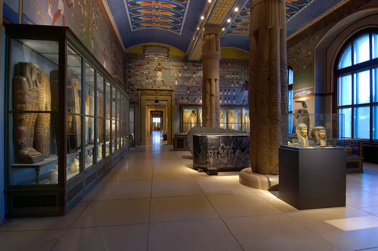 A spacious museum interior showcasing ancient Egyptian artifacts, including glass display cases with mummies, sculptures, and hieroglyphs along the walls. The ceiling features vibrant blue and gold artwork, and tall columns are visible throughout the room, creating a grand atmosphere.