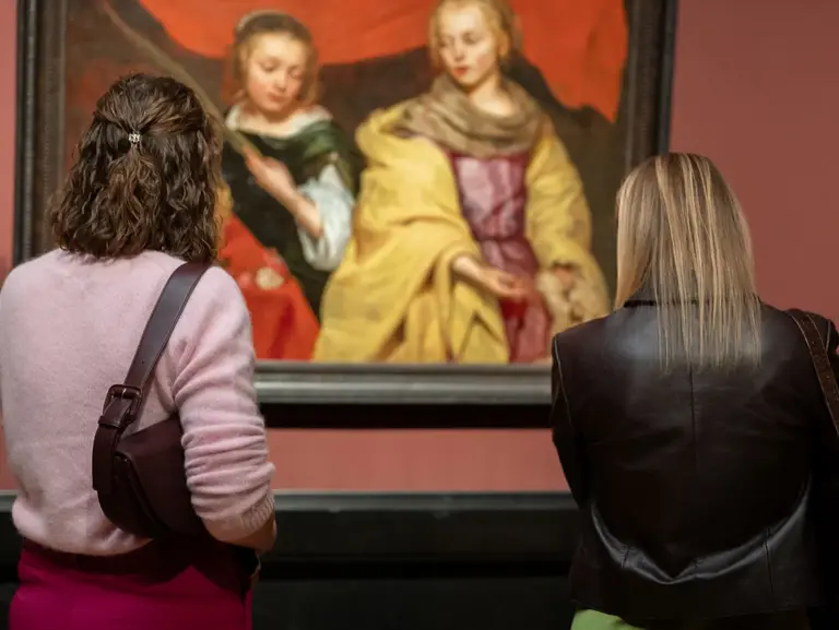 Two women stand in an art gallery, admiring a painting of two figures. The artwork features soft colors and flowing drapery, capturing the attention of the viewers as they engage with the art piece. A label is visible below the painting, providing information.