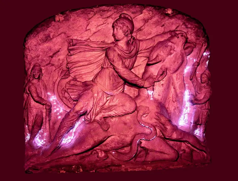 Eine reliefartige Darstellung zeigt einen mythologischen Helden, der auf einem Tier sitzt und es mit einer Hand hält. Im Hintergrund sind weitere Figuren angedeutet. Das Bild ist in einem rötlichen Licht erhellt, wodurch die Details und Texturen der Skulptur hervorgehoben werden.