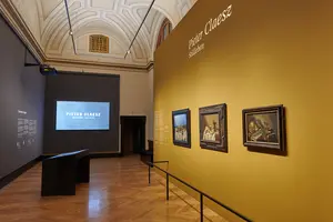 Das Bild zeigt eine Ausstellung über den Maler Pieter Claesz. An einer Wand sind Gemälde mit Stillleben zu sehen, während auf einer Leinwand im Hintergrund Informationen über den Künstler präsentiert werden. Der Bereich hat einen eleganten Holzfußboden und hohe Decken.