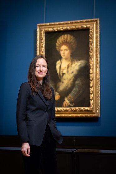 Eine Frau mit langen, braunen Haaren in einem schwarzen Blazer steht neben einem Gemälde in einem goldenen Rahmen. Das Bild im Hintergrund zeigt eine Frau mit auffälligem Kopfschmuck und einem eleganten Gewand. Die Wand ist blau gestrichen.