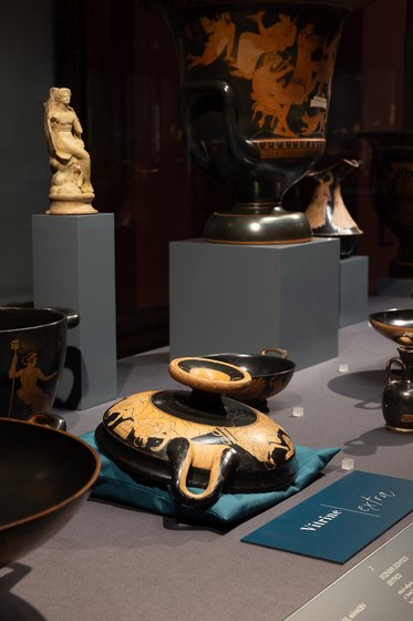 Alterntativtext: Eine Ausstellung von antiken griechischen Vasen und Töpferwaren, darunter ein Schalenobjekt mit dekorativem Muster, eine Figur auf einem Sockel und mehrere andere Keramiken. Die Objekte sind auf einem Tisch mit grauer Tischdecke angeordnet.