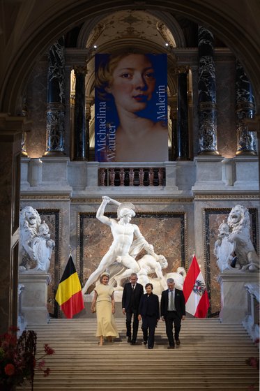 Eine elegante Gruppe von Menschen steht auf einer Treppe in einem prächtigen Raum. Im Hintergrund ist ein großes Banner mit dem Bild einer Frau und der Aufschrift „Madeleine“. Links und rechts der Treppe befinden sich Flaggen von Belgien und Österreich sowie beeindruckende Skulpturen.
