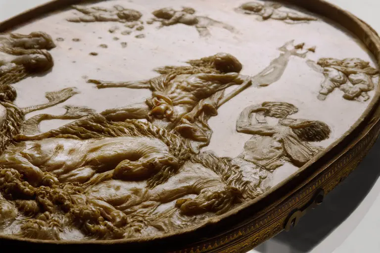 Eine ovalförmige Reliefdarstellung aus Elfenbein zeigt eine mythologische Szene mit verschiedenen Figuren. Im Vordergrund ist ein gefallener Krieger sichtbar, während andere Figuren, darunter kämpfende Gestalten und Tiere, um ihn herum arrangiert sind. Die Details des Reliefs sind tief und kunstvoll herausgearbeitet.
