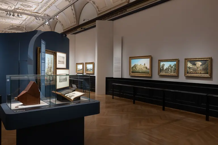 Das Bild zeigt einen Ausstellungsraum in einem Museum mit verschiedenen gerahmten Gemälden an der Wand. Im Vordergrund befindet sich eine Vitrine mit einem Modell und einem Buch. Der Raum hat hohe Decken und elegante architektonische Details.