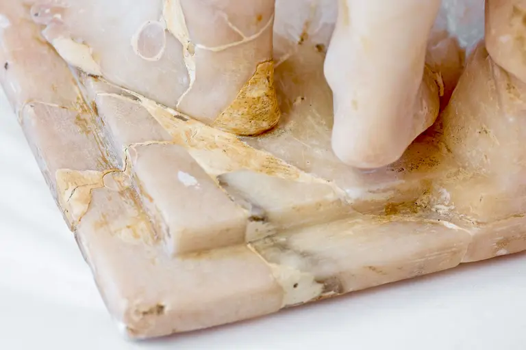 Eine Nahaufnahme einer Skulptur aus Alabaster zeigt detaillierte Texturen und Farbnuancen des Materials. Die Basis der Skulptur ist sichtbar und weist Risse sowie verschiedene Farbtöne auf, die die natürliche Beschaffenheit des Alabasters hervorheben.