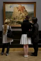 Drei Besucher stehen vor einem Gemälde, das den Turm zu Babel darstellt. Die monumentale Struktur ist von zahlreichen Gebäuden umgeben, und die Menschen im Bild scheinen über das Kunstwerk zu diskutieren.