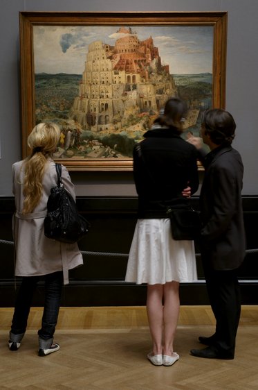 Drei Besucher stehen vor einem Gemälde, das den Turm zu Babel darstellt. Die monumentale Struktur ist von zahlreichen Gebäuden umgeben, und die Menschen im Bild scheinen über das Kunstwerk zu diskutieren.