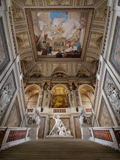 Die Aufnahme zeigt eine prachtvolle Treppe in einem historischen Gebäude mit aufwendigen Wandmalereien und Skulpturen. Im Hintergrund ist eine kunstvolle Decke mit Stuckverzierungen zu sehen. Die Atmosphäre strahlt Eleganz und Geschichte aus.