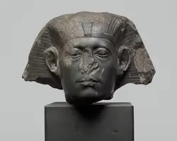 Eine antike Statue eines königlichen Kopfes mit stark abgerundeten Gesichtszügen und einer auffälligen Kopfdeckung. Der Kopf ist aus schwarzem Stein geschnitzt und zeigt einige Beschädigungen. Die Statue steht auf einem rechteckigen Sockel ohne weitere Dekoration.