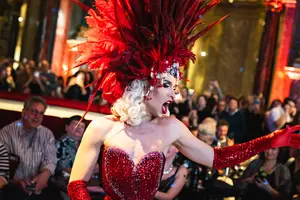Eine Drag-Künstlerin in einem glitzernden, roten kostümierten Outfit mit auffälligem Federkopfschmuck präsentiert sich auf einer Bühne. Im Hintergrund ist ein Publikum zu sehen, das die Darbietung begeistert verfolgt. Die Atmosphäre ist festlich und lebhaft.