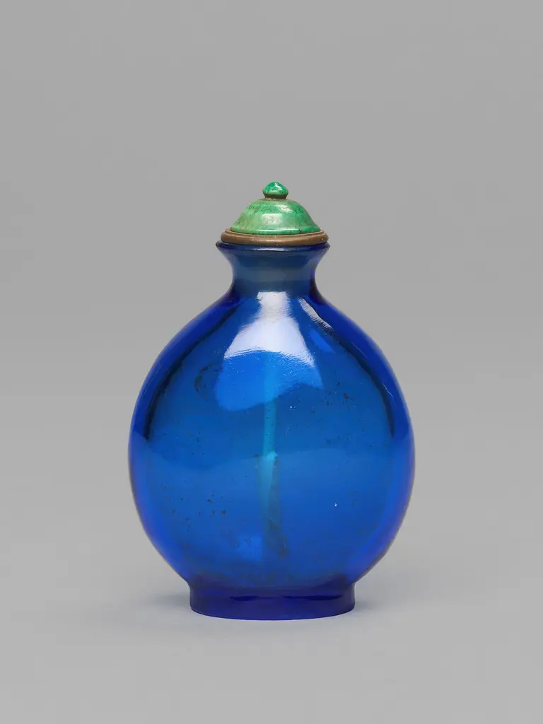 Eine ovale, blaue Glasflasche mit einer grünen Kappe. Der Flaschenhals ist schmal, und die Flasche hat eine glänzende Oberfläche, die das Licht reflektiert. Der Verschluss ist rund mit einem kleinen, grünen Kugelaufsatz.