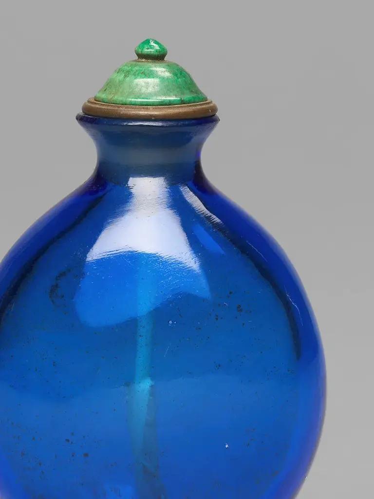 Eine ovale, blaue Glasflasche mit einer grünen Kappe. Der Flaschenhals ist schmal, und die Flasche hat eine glänzende Oberfläche, die das Licht reflektiert. Der Verschluss ist rund mit einem kleinen, grünen Kugelaufsatz.