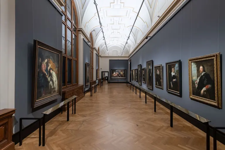Eine moderne Kunstgalerie mit hohen Decken und eleganten Details. An den Wänden hängen mehrere Gemälde in verschiedenen Rahmen, während im Hintergrund ein weiteres großes Gemälde zu sehen ist. Der Boden aus Holz und die Bänke schaffen eine einladende Atmosphäre für Besucher.
