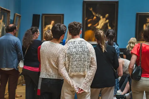A group of people stands in the Picture Gallery observing several artworks.