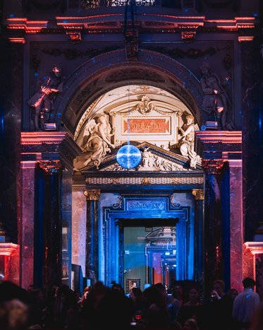 Ein prunkvoller Eingangsbereich mit einer großen, beleuchteten Decke und einer Discokugel. Die Wände sind mit kunstvollen Verzierungen und Statuen geschmückt, während das Licht in roten und blauen Tönen schimmert. Viele Menschen sind im Raum.