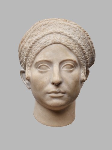 Eine antike Skulptur, die den Kopf einer Frau darstellt. Sie hat einen aufwendigen Haarknoten und einen ernsten Ausdruck. Die Statue ist aus hellem Stein gefertigt und hat detaillierte Gesichtszüge, darunter geschlossene Augen und eine weich konturierte Haut. Der Hintergrund ist neutral.