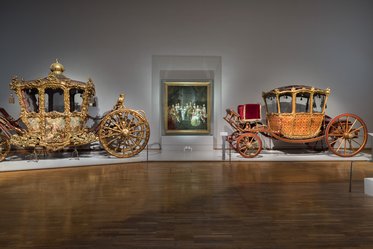 Zwei prächtig verzierte Kutschen stehen in einem Ausstellungsraum. Links eine goldene Kutsche mit aufwändigen Verzierungen, rechts eine rot-goldene Kutsche mit einem eleganten Sitz. Im Hintergrund ist ein Gemälde mit historischen Figuren sichtbar.