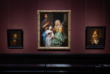 Ein Gemälde zeigt eine biblische Szene mit einer Mutter, die ein Kind hält, umgeben von zwei weiteren Figuren. Links und rechts hängen kleinere Bilder von Männern mit nachdenklichen Gesichtsausdrücken. Die Wände sind in einem tiefen Rot gestrichen, was die Kunstwerke hervorhebt.