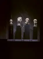 Vier antike Büsten aus Marmor stehen in einem dunklen Ausstellungsraum. Jede Büste ist auf einem schwarzen Sockel platziert und hebt sich vor der grauen Wand ab. Die Gesichter zeigen verschiedene Ausdrücke und Haarschnitte, was auf unterschiedliche historische Figuren hinweist.