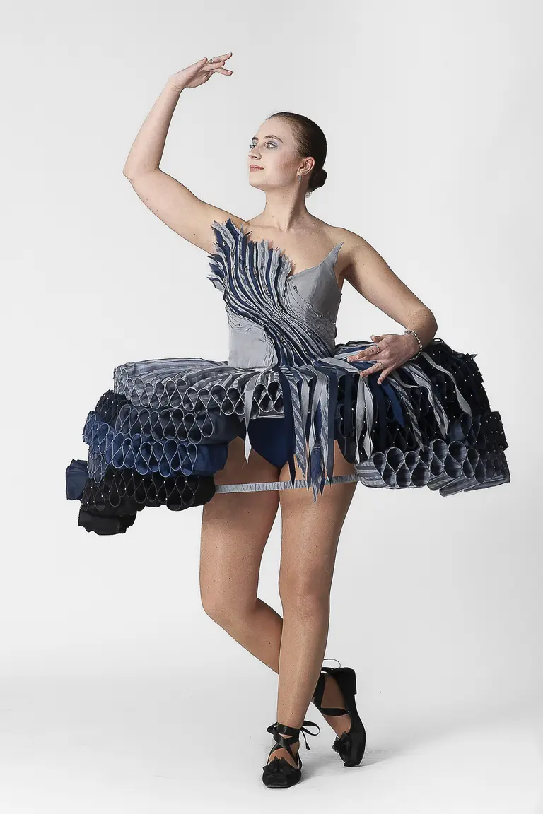 A model poses gracefully in a unique dress made of layered, folded fabric in shades of blue and gray. The dress has a structured, voluminous skirt and a fitted bodice, complimented by ballet flats. The background is plain, highlighting the design and the model's expressive stance.