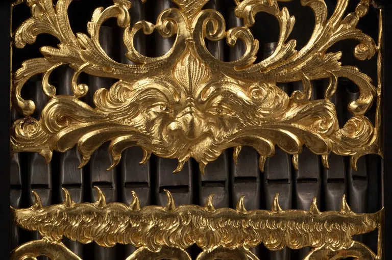 Das Bild zeigt kunstvoll gegossene, verzierte goldene Ornamente mit komplexen Mustern und Gesichtern, die üppige Details aufweisen. Die Elemente sind auf einer dunklen Oberfläche angeordnet und vermitteln einen Eindruck von Eleganz und Handwerkskunst.