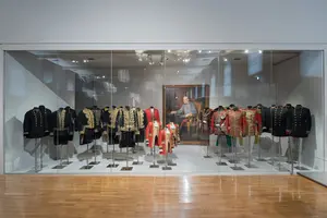 Im Bild ist ein Vitrinenausstellungsraum zu sehen, in dem eine Reihe historischer Uniformen präsentiert wird. Die Uniformen sind in verschiedenen Farben und Stilen, hauptsächlich schwarz mit goldenen Verzierungen, angeordnet. Im Hintergrund hängt ein Porträt einer historischen Figur.