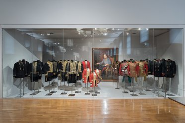 Im Bild ist ein Vitrinenausstellungsraum zu sehen, in dem eine Reihe historischer Uniformen präsentiert wird. Die Uniformen sind in verschiedenen Farben und Stilen, hauptsächlich schwarz mit goldenen Verzierungen, angeordnet. Im Hintergrund hängt ein Porträt einer historischen Figur.