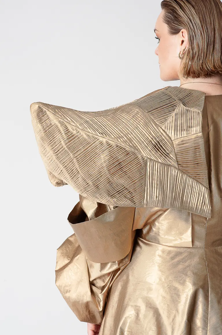 A model is seen from the side, showcasing a unique gold dress with an oversized, textured sleeve featuring angular patterns. The garment has a modern, architectural design, and the model’s hair is styled in a sleek, contemporary look.