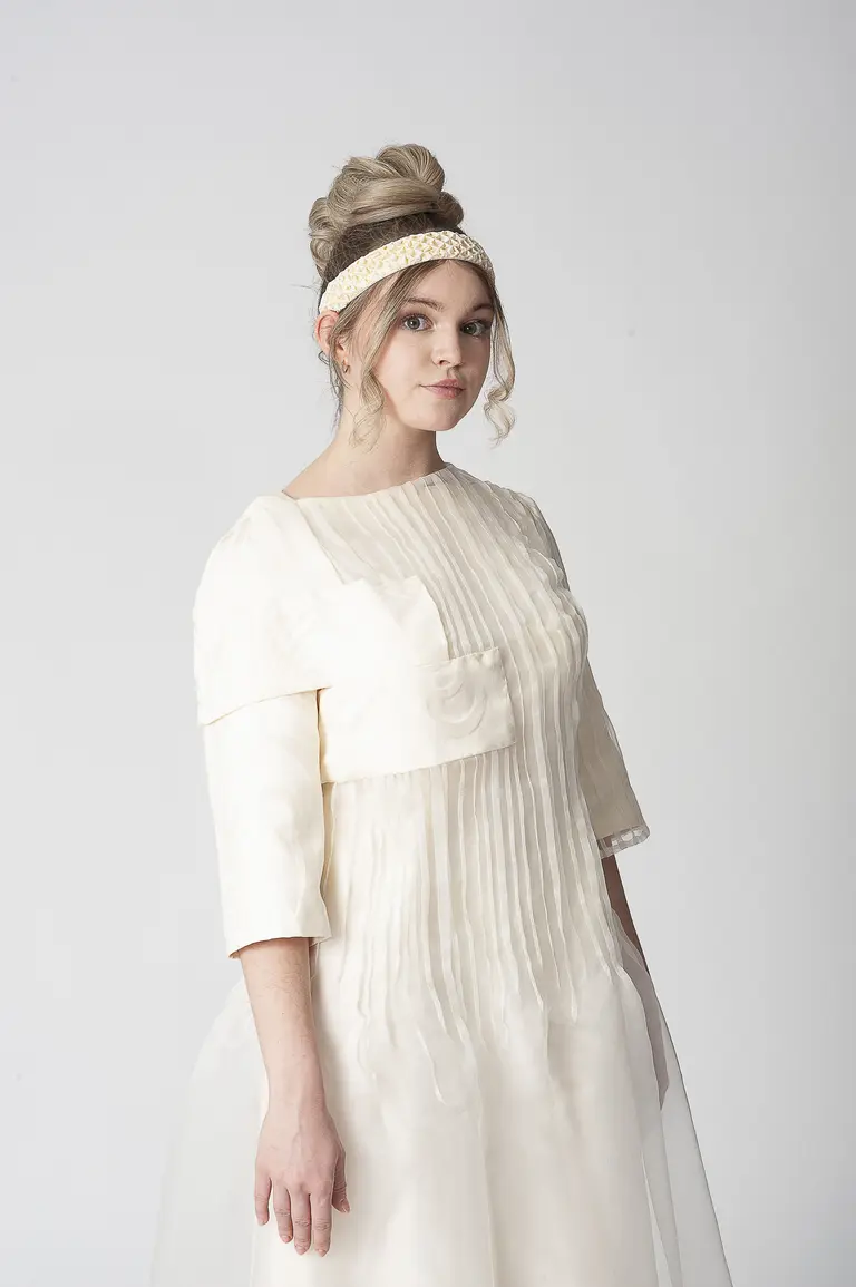 A model stands facing forward, wearing a light-colored, pleated dress with three-quarter sleeves and a flowing skirt. She has a stylish updo and is adorned with a decorative headband. The background is plain white, highlighting her elegant attire.