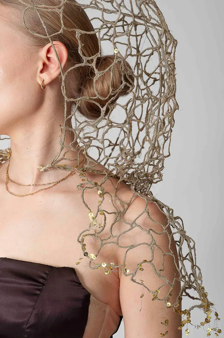 Close-up of a person wearing a unique, intricate golden net-like accessory draped over one shoulder. The accessory features delicate embellishments that catch the light, adding a touch of elegance to the outfit. The background is neutral, emphasizing the fashion detail.
