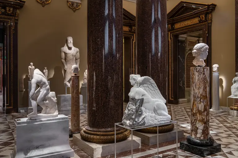 Eine Kunstgalerie mit mehreren Skulpturen aus Marmor und Stein. Zu sehen sind eine menschliche Figur, eine geflügelte sphinxähnliche Statue und verschiedene Büsten. Die Architektur umfasst hohe Säulen und dekorative Elemente, die dem Raum eine klassische Atmosphäre verleihen.