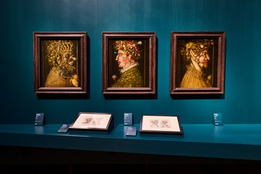 Drei gerahmte Porträts, die profilierte Gesichter zeigen, sind an einer teal-farbenen Wand aufgehängt. Jedes Bild hat eine unterschiedliche Figur, dekoriert mit verschiedenen Pflanzen und Früchten. Vor den Bildern liegen Zeichnungen auf einem Tisch, begleitet von Informationskarten.