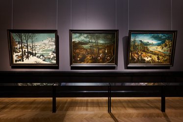 Drei Gemälde aus der Kunstgeschichte hängen an der Wand einer Galerie, dargestellt in eleganten Holzrahmen. Das erste Bild zeigt eine Winterlandschaft, das zweite ein bewaldetes Gebiet und das dritte eine idyllische Szene mit Tieren und einer hügeligen Landschaft.