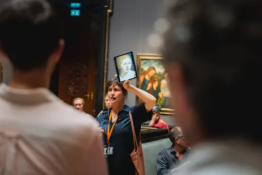 A museum guide stands in the Picture Gallery holding a tablet displaying a painting, engaging a group of visitors. The setting features paintings on the wall, with attendees listening attentively.