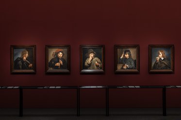 Fünf Gemälde in eleganten Rahmen an einer rot gestrichenen Wand. Jedes Bild zeigt eine einzelne Person, die verschiedene Ausdrücke und Haltungen zeigt. Im Vordergrund befinden sich dunkle Tische, die stationär sind. Die Atmosphäre ist künstlerisch und fördernd.