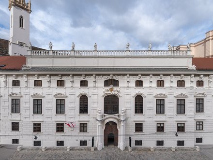Das Bild zeigt die Fassade eines weißen historischen Gebäudes mit vielen Fenstern und einer prächtigen Eingangstür. Über dem Eingang befindet sich ein Wappen. Im Hintergrund sind einige Skulpturen auf dem Dach sichtbar und der Himmel ist teilweise bewölkt.