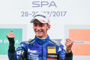 Ein junger Rennfahrer in einem blauen Rennanzug und einer weißen Kappe hält seine Hände in einer Siegerpose. Er lächelt freudig, während er vor einem hinteren Banner mit dem Text „SPA 28-30/07/2017“ steht.