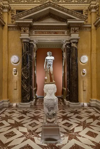 Die Abbildung zeigt eine Statue von David in einem Museum. Sie steht majestätisch im Zentrum eines Raumes, umgeben von Säulen und kunstvollen Wänden. Der Boden ist mit einem geometrischen Muster gestaltet, und es sind zarte Reliefs an den Wänden zu sehen.
