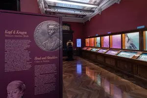 Die Ausstellung zeigt eine historische Modekollektion mit Fokus auf Kopf- und Kragenmode für Männer. Im Vordergrund steht ein Info-Tafel mit Texten zu den Themen. Im Hintergrund sind Vitrinen mit weiteren Exponaten sowie farbige Beleuchtung, die die Mode präsentiert.