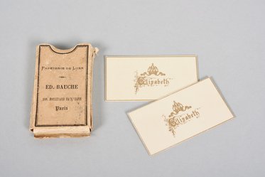 Ein kleines, älteres Notizbuch mit dem Titel "Papeterie de Luxe" von ED. BAUCHE in Paris steht links. Daneben liegen zwei elegante, cremefarbene Visitenkarten mit dem Namen "Elizabeth" in goldenen Buchstaben.