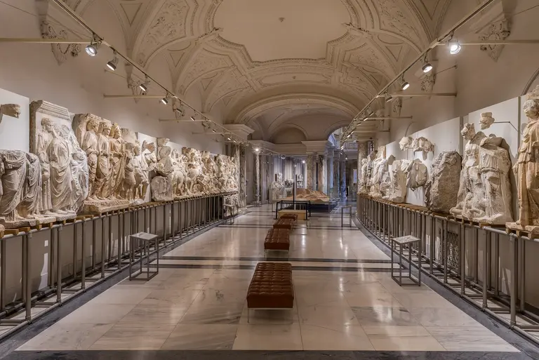 Ein langer, heller Ausstellungsraum mit antiken Marmorreliefs, die an den Wänden präsentiert werden. In der Mitte stehen Sitzbänke, und die Beleuchtung hebt die Details der Skulpturen hervor. Der Raum strahlt eine ruhige, kulturelle Atmosphäre aus.